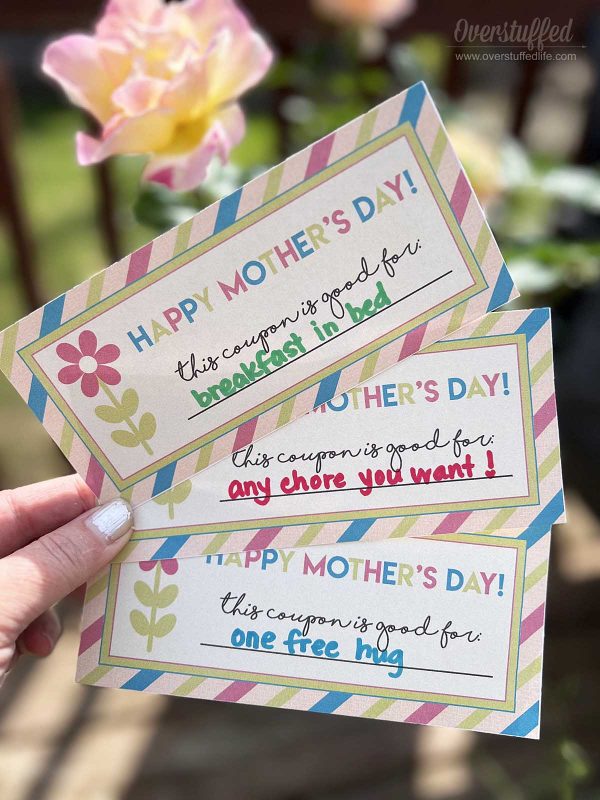 Mother's Day coupons held in front of a rose. Coupons say breakfast in bed, any chore you want, and one free hug